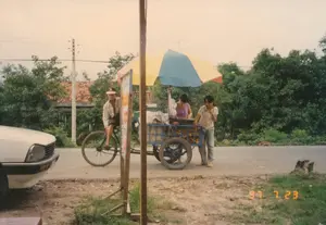 Bức ảnh chụp cảnh sinh hoạt ven đường ở một vùng quê Việt Nam, có thể là ở khu vực Củ Chi hoặc ngoại ô Sài Gòn, vào ngày 23 tháng 7 năm 1997, như được ghi nhận bởi dấu ngày tháng '97 7 23' ở góc dưới bên phải.。Địa điểm là một con đường nhựa. Ở trung tâm bức ảnh là một quầy hàng rong trên chiếc xe lôi (xe ba gác đạp). Chiếc xe có khung màu xanh lam, chở đầy các sọt trái cây màu cam, có thể là cam hoặc quýt. Một cây dù lớn nhiều màu sắc (vàng, xanh, đỏ) được cắm trên xe để che nắng.。Hoạt động chính là cảnh buôn bán. Có ba người trong ảnh: một người đàn ông đội mũ lưỡi trai đang ngồi trên phần xe đạp của chiếc xe lôi; một người phụ nữ (người bán) đang đứng sau quầy hàng; và một người phụ nữ khác (khách hàng) đang đứng phía trước, dường như đang lựa chọn hoặc ăn thử trái cây.。Ở phía trước, có một tấm biển quảng cáo dựng trên khung gỗ nhưng chữ viết trên đó bị mờ và không thể đọc được. Bên trái ảnh là một phần đầu của một chiếc ô tô cũ màu trắng. Phông nền là cây cối rậm rạp, một cột điện và thấp thoáng một ngôi nhà mái ngói đỏ.。（日本語訳）。右下隅にある「'97 7 23」という日付スタンプに記録されているように、1997年7月23日に、ベトナムの田舎、おそらくクチ地区かサイゴン郊外で撮影された道端の生活風景の写真です。場所はアスファルトの道路です。写真の中央には、シクロ（三輪自転車）の上の露店があります。その車両は青いフレームで、オレンジ色の果物、おそらくオレンジかみかんが入ったカゴでいっぱいです。日差しを避けるために、大きな多色（黄色、青、赤）のパラソルが車両に立てられています。主な活動は商売の光景です。写真には3人の人物がいます。キャップをかぶった男性がシクロの自転車部分に座っており、一人の女性（売り手）が店の後ろに立ち、そしてもう一人の女性（客）が前に立って、果物を選んでいるか試食しているようです。前景には、木製のフレームに立てられた広告看板がありますが、その上の文字はぼやけていて読むことができません。写真の左側には、古い白い車の前部が見えます。