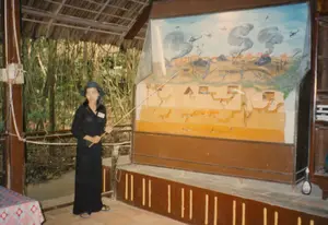 Bối cảnh là một khu trưng bày tại di tích lịch sử Địa đạo Củ Chi. Đây là một không gian bán ngoài trời có mái lợp lá, cột gỗ và nền lát gạch, phía sau là rừng cây. Một nữ hướng dẫn viên mặc trang phục màu đen và đội nón đang thuyết minh. Cô dùng một que chỉ để giới thiệu về một sa bàn lớn. Sa bàn này mô tả chi tiết cấu trúc mặt cắt của hệ thống địa đạo với nhiều tầng hầm phức tạp. Phần trên của sa bàn tái hiện một cảnh chiến đấu trên mặt đất trong thời chiến, bao gồm máy bay trực thăng, xe tăng, và các cột khói từ những vụ nổ. Trên ngực áo của người phụ nữ có một bảng tên nhưng không thể đọc được chữ. Ở phía bên trái có một chiếc bàn được phủ khăn trải bàn.。（日本語訳）。背景は、クチの地下道歴史遺跡地区にある展示エリアです。ここは、葉葺きの屋根、木製の柱、タイル張りの床を持つ半屋外の空間で、後ろには林があります。黒い服を着てノンラーを被った女性ガイドが説明をしています。彼女は指し棒を使って、大きなジオラマについて紹介しています。このジオラマは、多くの複雑な地下階層を持つ地下道システムの断面構造を詳細に描写しています。ジオラマの上部は、ヘリコプター、戦車、そして爆発による煙の柱など、戦時中の地上での戦闘シーンを再現しています。女性の胸には名札がありますが、文字は読み取れません。左側には、テーブルクロスで覆われたテーブルがあります。