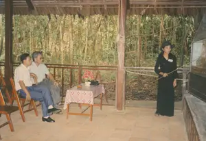 Bối cảnh là một khu vực thuyết minh ngoài trời có mái che lợp lá, cột gỗ và sàn lát gạch, có khả năng là tại một khu di tích lịch sử như Địa đạo Củ Chi. Phía sau là một khu rừng rậm rạp với nhiều cây tre.。Một người phụ nữ, có vẻ là hướng dẫn viên, mặc trang phục màu đen, đội nón và đeo một bảng tên (chữ không đọc được), đang đứng thuyết trình. Cô dùng một cây que dài để chỉ vào một mô hình hoặc sa bàn được đặt trong tủ kính lớn.。Hai người đàn ông đang ngồi trên ghế xếp bằng gỗ để lắng nghe. Người đàn ông ngồi phía trước mặc áo sơ mi trắng tay ngắn, quần tây xanh và đi giày. Người đàn ông ngồi sau mặc áo sơ mi sáng màu, quần tối màu, đeo kính râm và đi dép.。Ở giữa họ là một chiếc bàn gỗ nhỏ được phủ khăn trải bàn có hoa văn. Trên bàn có một lọ hoa giả màu đỏ và một cái gạt tàn thuốc.。（日本語訳）。背景は、葉葺きの屋根、木の柱、タイル張りの床がある屋外の説明エリアで、クチトンネルのような歴史遺跡地区である可能性があります。後ろには竹がたくさんある鬱蒼とした森があります。ガイドと思われる一人の女性が、黒い服を着て、帽子をかぶり、名札（文字は読めない）を付け、立って説明しています。彼女は長い棒を使って、大きなガラスケースの中に置かれた模型またはジオラマを指し示しています。二人の男性が木製の折りたたみ椅子に座って聞いています。前に座っている男性は白い半袖シャツ、青いズボンを履き、靴を履いています。後ろに座っている男性は明るい色のシャツ、暗い色のズボンを履き、サングラスをかけ、サンダルを履いています。彼らの間には、模様のあるテーブルクロスで覆われた小さな木製のテーブルがあります。テーブルの上には、赤い造花の花瓶と灰皿があります。