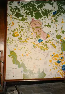 Bức ảnh chụp một tấm bản đồ lớn được đóng trong khung gỗ, có vẻ như đang được trưng bày trong một bảo tàng hoặc khu di tích lịch sử, có thể là tại khu vực Củ Chi. Sàn nhà được lát gạch tối màu.。Các thông tin văn bản có thể đọc được trên bản đồ bao gồm:。- Ở góc dưới bên trái: 'TỶ LỆ 1/25.000' và 'CHÚ THÍCH'. Phần chú thích giải nghĩa các ký hiệu trên bản đồ nhưng chữ rất mờ.。- Tên các địa danh trên bản đồ, dù một số bị mờ, bao gồm: 'xã Phú Mỹ Hưng', 'xã An Nhơn Tây', 'xã Nhuận Đức', 'xã An Phú', 'xã Trung Lập Thượng', 'xã Trung Lập Hạ'.。- Một khu vực lớn được tô màu hồng có các ký hiệu quân sự, có thể là khu căn cứ.。- Các từ chỉ địa giới như 'TỈNH' và 'HẬU' cũng xuất hiện.。Về quang cảnh và hoạt động:。- Địa điểm: Một không gian trong nhà, có khả năng là phòng trưng bày của một khu di tích.。- Hoạt động: Bức ảnh ghi lại một hiện vật tĩnh đang được trưng bày. Không có hoạt động nào của con người diễn ra.。- Vật thể: Đối tượng chính là tấm bản đồ quân sự, thể hiện địa hình với sông ngòi (màu xanh dương), rừng cây (màu xanh lá), các khu vực khác (màu vàng, hồng). Tấm bản đồ được đặt trong khung gỗ. Dưới sàn nhà có một vật thể trông giống như một phần của cái chổi hoặc vật dụng đan lát.。（日本語訳）。写真は、木製の額縁に収められた大きな地図を写したもので、博物館か史跡、おそらくクチ地区で展示されているようです。床は暗い色のタイル張りです。地図上で読み取れる文字情報は以下の通りです。- 左下隅：「縮尺 1/25,000」および「凡例」。凡例部分は地図上の記号を説明していますが、文字は非常に不鮮明です。- 地図上の地名には、一部不鮮明なものもありますが、「フーミーフン社」、「アンニョンタイ社」、「ニュアンドゥック社」、「アンフー社」、「チュンラップトゥオン社」、「チュンラップハ社」などが含まれます。- 軍事記号が記された広いピンク色のエリアがあり、基地である可能性があります。- 「TỈNH（省）」や「HẬU」といった境界を示す言葉も現れています。光景と活動について：。- 場所：屋内空間で、史跡の展示室である可能性があります。- 活動：写真は展示されている静的な展示物を記録したものです。人間の活動は行われていません。- 物体：主な対象は軍事地図で、河川（青色）、森林（緑色）、その他の地域（黄色、ピンク色）で地形を表しています。地図は木製の額縁に収められています。床には、ほうきの一部または編み物のような物体が見えます。