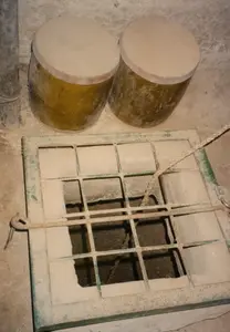 Bức ảnh chụp một cảnh bên trong một cấu trúc dưới lòng đất, có khả năng là một phần của Địa đạo Củ Chi. Ở phía trước là một miệng hố hình vuông được đậy bằng một tấm lưới sắt đã gỉ sét và bám đầy bụi. Tấm lưới có khung màu xanh lá cây đã cũ, một số thanh sắt đã bị gãy. Một sợi dây thừng dày được thả xuống hố qua tấm lưới. Phía sau miệng hố là hai cái chum lớn màu vàng, có nắp đậy, có thể được dùng để chứa nước hoặc lương thực. Toàn bộ không gian có sàn và tường bằng đất, gợi lên khung cảnh của một hầm trú ẩn hoặc khu vực sinh hoạt dưới lòng đất.。（日本語訳）。写真は地下構造物の内部の光景を写しており、クチトンネルの一部である可能性があります。手前には、錆びて埃をかぶった鉄格子で覆われた四角い穴があります。格子には古い緑色の枠があり、鉄の棒のいくつかは折れています。太いロープが格子を通って穴に下ろされています。穴の後ろには、蓋付きの大きな黄色い壺が二つあり、水や食料を貯蔵するために使われていた可能性があります。空間全体は土の床と壁でできており、防空壕や地下の生活区域の光景を思い起こさせます。