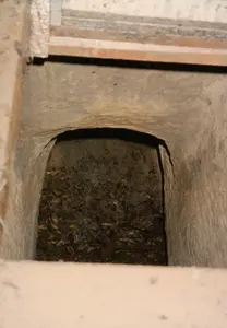 Hình ảnh chụp nhìn từ trên xuống một lối vào của đường hầm dưới lòng đất. Đây có khả năng là một phần của hệ thống Địa đạo Củ Chi, một di tích lịch sử. Lối vào là một cái hố hẹp, hình chữ nhật, có thành được làm bằng đất. Bên dưới là một đường hầm tối, nhỏ hẹp với phần nóc có dạng vòm. Dưới đáy hầm có thể thấy đất và nhiều lá cây khô, có thể được dùng để ngụy trang. Miệng hố được bao quanh bởi một cấu trúc trông giống như khung gỗ hoặc bê tông.。（日本語訳）。地下トンネルの入り口を上から見下ろした写真。これは歴史的遺跡であるクチトンネルシステムの一部である可能性があります。入り口は狭い長方形の穴で、壁は土でできています。その下には、天井がアーチ状になった、暗く狭いトンネルがあります。トンネルの底には土とたくさんの枯れ葉が見え、カモフラージュに使われていた可能性があります。穴の口は、木枠かコンクリートのような構造物で囲まれています。