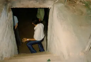 Bức ảnh ghi lại cảnh một nhóm người đang đi xuống một lối vào hẹp của một đường hầm dưới lòng đất, rất có thể là tại khu di tích lịch sử Địa đạo Củ Chi. Lối vào có cầu thang bằng bê tông dẫn xuống khu vực tối tăm bên dưới. Các bức tường xung quanh được làm bằng bê tông hoặc đất nén, tạo cảm giác kiên cố nhưng chật hẹp. Một người đàn ông mặc áo sơ mi trắng cộc tay và quần tây xanh sẫm đang khom người cẩn thận bước xuống. Phía trước anh ta, trong bóng tối, có thể thấy một phần của hai người khác, một người mặc áo xanh nhạt và một người mặc áo xanh lá cây. Hoạt động này là một chuyến tham quan, trải nghiệm không gian bên trong địa đạo. Ở góc dưới bên phải của bức ảnh có in dấu ngày tháng màu cam '97 7 23', xác nhận thời điểm chụp là ngày 23 tháng 7 năm 1997.。（日本語訳）。写真は、一団の人々が地下トンネルの狭い入り口を下りている光景を記録したもので、おそらく歴史的遺跡であるクチトンネルでのものでしょう。入り口には、下の暗いエリアへと続くコンクリート製の階段があります。周囲の壁はコンクリートまたは固められた土でできており、頑丈でありながらも狭苦しい感じを与えます。白い半袖シャツと濃い青色のズボンを履いた男性が、慎重に身をかがめて下りています。彼の前方、暗闇の中に、他の二人の一部が見えます。一人は水色のシャツを、もう一人は緑色のシャツを着ています。この活動は、トンネル内部の空間を体験する見学ツアーです。写真の右下隅にはオレンジ色の日付スタンプ「'97 7 23」が印字されており、撮影日時が1997年7月23日であることを示しています。