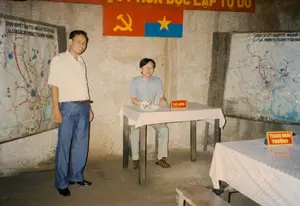 Các dòng chữ trong ảnh bao gồm:。- Khẩu hiệu trên tường: "[KHÔNG CÓ GÌ QUÝ] HƠN ĐỘC LẬP TỰ DO".。- Tiêu đề trên bản đồ bên trái: "SƠ ĐỒ QUYẾT TÂM TTC-NỔI DẬY TẾT MẬU THÂN 1968", "LL CỦA CÁC PK TRÊN CT SÀI GÒN-GIA ĐỊNH".。- Tiêu đề trên bản đồ bên phải: "SƠ ĐỒ QUYẾT TÂM TTC-NỔI DẬY... SÀI GÒN".。- Biển chức danh trên bàn: "TƯ LỆNH" và "THAM MƯU TRƯỞNG".。- Dấu ngày tháng trên khăn trải bàn: "97 7 23".。Bối cảnh là một căn phòng họp hoặc hầm chỉ huy quân sự được tái hiện, có khả năng nằm trong khu di tích lịch sử Địa đạo Củ Chi. Hai người đàn ông đang chụp ảnh kỷ niệm trong không gian này. Một người đàn ông mặc áo sơ mi và quần jean đang đứng, trong khi người còn lại đeo kính, ngồi tại chiếc bàn có biển chức danh "TƯ LỆNH". Trên tường treo cờ Đảng Cộng sản Việt Nam và cờ Mặt trận Dân tộc Giải phóng miền Nam Việt Nam, cùng với hai tấm bản đồ quân sự lớn về chiến dịch Tết Mậu Thân 1968 tại Sài Gòn. Các đồ vật như bàn ghế gỗ đơn sơ, bộ ấm chén trà và các biển chức danh được sắp đặt để mô phỏng một phòng chỉ huy thời chiến.。（日本語訳）。写真の中の文字は以下の通りです：。- 壁のスローガン：「独立と自由ほど[尊いものはない]」。- 左の地図のタイトル：「1968年テト攻勢における総攻撃・蜂起決意図」、「サイゴン-ジャディン戦線における各分区の戦力」。- 右の地図のタイトル：「総攻撃・蜂起決意図...サイゴン」。- 机の上の役職プレート：「司令官」と「参謀長」。- テーブルクロスの日付：「97 7 23」。背景は、再現された会議室または軍事司令部壕であり、クチトンネル歴史地区内にある可能性があります。二人の男性がこの空間で記念撮影をしています。一人の男性はシャツとジーンズを着用して立っており、もう一人の眼鏡をかけた男性は「司令官」の役職プレートがある机に座っています。壁にはベトナム共産党旗と南ベトナム解放民族戦線旗が、サイゴンでの1968年テト攻勢に関する2枚の大きな軍事地図と共に掲げられています。簡素な木製の机と椅子、茶器セット、役職プレートなどの物品が、戦時中の司令室を模して配置されています。