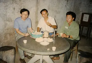 Bối cảnh của bức ảnh là một không gian trong nhà với những bức tường xi măng thô ráp, có khả năng là một phần của một đường hầm hoặc hầm trú ẩn, gợi nhớ đến không gian bên trong Địa đạo Củ Chi. Ba người đàn ông đang ngồi quanh một chiếc bàn tròn bằng đá. Họ ngồi trên những chiếc ghế đẩu làm từ gỗ và những khúc cây. Hoạt động chính là ba người đang nghỉ ngơi, ăn uống và trò chuyện. Trên bàn có một tô lớn màu xanh lam đựng khoai mì (sắn) luộc, các đĩa nhỏ hơn cũng đựng khoai mì đã cắt miếng, một chén nhỏ có thể là muối mè để chấm, và nhiều tách trà nhỏ. Người đàn ông ở giữa mặc áo sơ mi trắng đang cầm một miếng khoai mì. Người đàn ông bên phải mặc áo sơ mi xanh lá cây, có đeo bảng tên, đang cầm một tách trà. Ở phía sau bên phải, có một thùng quyên góp bằng gỗ đặt trên giá đỡ. Trên thùng có một tấm biển ghi chữ: 'ĐÓNG GÓP', 'DONATION', và các ký tự chữ Hán bên dưới.。（日本語訳）。写真の背景は、粗いセメントの壁がある屋内の空間で、トンネルか防空壕の一部である可能性があり、クチトンネルの内部空間を彷彿とさせます。3人の男性が石の丸テーブルを囲んで座っています。彼らは木や丸太で作られたスツールに座っています。主な活動は、3人が休憩し、食事をし、おしゃべりをしていることです。テーブルの上には、茹でたキャッサバ（タピオカ）が入った大きな青いボウル、同じくカットされたキャッサバが入った小さな皿、つけダレ用のゴマ塩と思われる小さな椀、そしてたくさんの小さな湯呑みがあります。中央の男性は白いシャツを着ており、キャッサバを一切れ持っています。右側の男性は緑色のシャツを着て名札を付けており、湯呑みを持っています。右奥には、台の上に置かれた木製の募金箱があります。箱には「ĐÓNG GÓP」、「DONATION」、そしてその下に漢字が書かれた札が付いています。