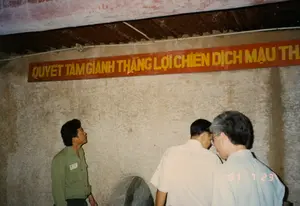 Trên tường có treo một biểu ngữ dài màu đỏ với dòng chữ vàng: 'QUYẾT TÂM GIÀNH THẮNG LỢI CHIẾN DỊCH MẬU THÂN'. Bối cảnh là bên trong một đường hầm hoặc hầm trú ẩn dưới lòng đất, có khả năng là tại khu di tích lịch sử Địa đạo Củ Chi. Các bức tường bằng đất thô sơ và trần nhà thấp được gia cố bằng các thanh gỗ và tấm tôn. Một người đàn ông mặc áo sơ mi màu xanh lá cây, có vẻ là hướng dẫn viên, đang nhìn lên biểu ngữ và thuyết minh cho hai du khách mặc áo sơ mi sáng màu. Ở phía dưới, có thể thấy một phần của một vật thể bằng đá, trông giống như một chiếc cối xay đá, một vật dụng sinh hoạt được trưng bày trong các di tích lịch sử.。（日本語訳）。壁には、「マウタン作戦の勝利を勝ち取る決意」という黄色い文字が書かれた長い赤い横断幕が掲げられています。背景は地下のトンネルまたは防空壕の中で、おそらくクチの地下道歴史遺跡地区でしょう。粗末な土壁と低い天井は、木材とトタン板で補強されています。緑色のシャツを着た男性はガイドのようで、横断幕を見上げ、明るい色のシャツを着た2人の観光客に説明しています。下の方には、石でできた物体の一部が見えます。それは石臼のように見え、史跡で展示されている生活用品です。