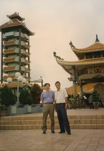 Bức ảnh chụp hai người đàn ông đang đứng tạo dáng chụp ảnh kỷ niệm tại một khu vực có kiến trúc giống đền chùa hoặc khu tưởng niệm. Dựa trên bối cảnh địa lý, đây có khả năng là Đền tưởng niệm liệt sĩ Bến Dược tại Củ Chi, được chụp vào năm 1997.。Phía sau hai người, bên trái là một ngọn tháp cao nhiều tầng với các tầng mái lợp ngói màu cam. Bên phải là một công trình lớn có kiến trúc cổ, có thể là cổng tam quan hoặc nhà bia, với phần mái cong lợp ngói âm dương và các đầu đao được trang trí cầu kỳ. Trên các cột trụ của công trình này có thể thấy các ký tự chữ viết nhưng không thể đọc rõ nội dung.。Hai người đàn ông đứng trên một khoảng sân rộng được lát gạch, phía sau họ có vài bậc tam cấp. Người đàn ông bên trái mặc áo phông màu tím và quần tây màu be. Người đàn ông bên phải mặc áo sơ mi trắng cộc tay và quần tây màu xanh đậm. Xung quanh khu vực có các bồn cây cảnh và cột đèn chiếu sáng công cộng hình quả cầu. Ở phía xa có vài người khác đang đi lại, cho thấy đây là một địa điểm tham quan.。（日本語訳）。この写真は、寺院または記念区域のような建築様式を持つ場所で、記念写真を撮るためにポーズをとって立っている2人の男性を写したものです。地理的背景に基づくと、これは1997年に撮影された、クチにあるベンズオック烈士記念祠である可能性があります。二人の後ろ、左側には、オレンジ色の瓦で葺かれた各層の屋根を持つ、高い多層の塔があります。右側には、古い建築様式の大きな建造物があり、それは三門か石碑堂である可能性があります。湾曲した屋根は陰陽瓦で葺かれ、軒先の飾りは精巧に装飾されています。二人の男性は煉瓦が敷かれた広い中庭に立っており、彼らの後ろには数段の階段があります。左側の男性は紫色のTシャツとベージュ色のズボンを着用しています。右側の男性は白い半袖シャツと濃い青色のズボンを着用しています。周辺には観賞植物の植木鉢や球形の公共照明灯があります。遠くには他の数人が歩き回っており、ここが観光地であることを示しています。