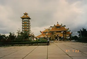 Trên cổng tam quan có dòng chữ 'ĐỀN BẾN DƯỢC'. Hai bên cột của cổng có treo các câu đối chữ dọc nhưng không thể đọc rõ nội dung. Đây là khung cảnh tại khu di tích lịch sử Đền Tưởng niệm Liệt sĩ Bến Dược ở Củ Chi, được chụp vào ngày 23 tháng 7 năm 1997. Công trình có kiến trúc truyền thống Việt Nam. Bên trái là một ngọn tháp tưởng niệm cao nhiều tầng với mái ngói màu cam. Ở trung tâm là cổng tam quan bề thế dẫn vào khu đền chính. Các mái nhà đều được lợp ngói và có đầu đao cong vút. Phía trước là một khoảng sân rộng lát gạch bê tông. Có một vài du khách đang tham quan khu di tích; hai người đang đứng ở sân, trong đó có một người đội nón lá, và một vài người khác đang đi về phía cổng chính. Quốc kỳ Việt Nam đang tung bay trên cột cờ. Xung quanh khu đền được trồng nhiều cây xanh, cây cảnh và có các cột đèn chiếu sáng. Bầu trời u ám, nhiều mây.。（日本語訳）。三門の上には「ĐỀN BẾN DƯỢC」という文字がある。門の両側の柱には縦書きの対句が掛けられているが、内容ははっきりと読み取れない。これは、1997年7月23日に撮影された、クチにあるベンズオック烈士記念祠歴史遺跡地区の光景である。この建造物はベトナムの伝統的な建築様式である。左側には、オレンジ色の瓦屋根を持つ多層の高い記念塔がある。中央には、本殿エリアへと続く壮大な三門がある。屋根はすべて瓦で葺かれ、軒先は反り上がっている。前方にはコンクリートタイルで舗装された広い中庭がある。数人の観光客が遺跡地区を見学している。2人が中庭に立っており、そのうちの1人はノンラーをかぶっている。そして、他の数人が正門の方へ向かって歩いている。ベトナムの国旗が旗竿にはためいている。祠の周りには多くの緑樹や観賞植物が植えられており、街灯の柱もある。空は曇っており、雲が多い。