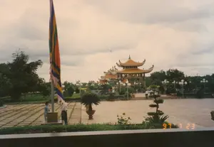Bức ảnh chụp một không gian rộng lớn, có khả năng là một khu tưởng niệm hoặc đền thờ tại Củ Chi, vào một ngày trời nhiều mây. Ở phía xa là các công trình kiến trúc mang phong cách truyền thống với mái ngói cong màu cam, có thể là cổng hoặc các nhà tưởng niệm. Một quảng trường rộng được lát gạch theo ô vuông chiếm phần lớn không gian. Ở phía trước bên trái, một lá cờ ngũ sắc lớn, giống cờ Phật giáo, đang được treo trên một cột cờ cao. Có hai người đang đứng gần cột cờ, một người dường như đang điều chỉnh lá cờ. Vài người khác đang đi bộ ở phía xa, gần các công trình kiến trúc. Xung quanh quảng trường được trang trí bằng nhiều cây xanh, cây cảnh bonsai, cây cọ trồng trong chậu lớn và các cột đèn chiếu sáng. Dấu ngày tháng "97 7 23" được hiển thị ở góc dưới bên phải của bức ảnh.。（日本語訳）。写真は、曇りの日に、クチにある記念地区または寺院と思われる広大な空間を写しています。遠方には、オレンジ色の反った瓦屋根を持つ伝統的な様式の建築物があり、門または記念館である可能性があります。広い広場が四角いタイルで舗装されており、空間の大部分を占めています。左手前には、仏旗に似た大きな五色の旗が高い旗竿に掲げられています。旗竿の近くに二人の人物が立っており、一人は旗を調整しているようです。他に数人が遠くの建築物の近くを歩いています。広場の周りは、多くの緑の木々、盆栽、大きな鉢に植えられたヤシの木、そして街灯柱で装飾されています。写真の右下の角に「97 7 23」という日付が表示されています。