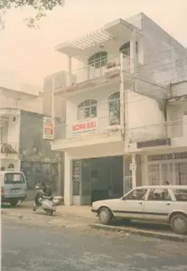 Bối cảnh là một con đường ở một thị trấn hoặc thành phố của Việt Nam vào năm 1997. Ở trung tâm là một tòa nhà nhiều tầng màu trắng, là một khách sạn mini. Có nhiều biển hiệu ghi rõ thông tin:。1.  Biển hiệu dọc bên trái: "MINI HOTEL HONG HAI", bên dưới có ghi địa chỉ "40* Bùi Thị Xuân" và số điện thoại "ĐT: 629990".。2.  Băng rôn lớn ở tầng hai: Chữ đỏ "HỒNG HẢI". Bên dưới là dòng chữ nhỏ hơn "Lịch sự Cho đáo Đón se trong nhở" (có thể là biến thể của "Lịch sự, Chu đáo, Đón tiếp trọng thị"), cùng với địa chỉ và số điện thoại.。3.  Bảng đen ở lối vào: "CAFE", "Giải khát", "KARAOKE", cho thấy nơi đây cũng kinh doanh dịch vụ ăn uống và giải trí.。4.  Biển treo nhỏ bên phải: "cho thuê truyện".。Về hoạt động và vật thể, một người đàn ông đang ngồi trên chiếc xe tay ga kiểu Vespa cũ đậu bên lề đường. Gần đó có một vài bao tải. Một chiếc ô tô kiểu station wagon màu trắng (giống xe Peugeot 505) đang đậu ở phía bên phải. Xa hơn một chút về bên trái là một chiếc xe van. Con đường được trải nhựa nhưng có vẻ đã cũ. Hệ thống dây điện chằng chịt vắt ngang qua mặt tiền của các tòa nhà, một hình ảnh đặc trưng của đô thị Việt Nam thời kỳ này.。（日本語訳）。背景は1997年のベトナムのある町、または都市の通りです。中央には白い多層階の建物があり、それはミニホテルです。情報を明記した多くの看板があります：。1.  左側の縦長の看板：「MINI HOTEL HONG HAI」、その下には住所「40* Bùi Thị Xuân」と電話番号「ĐT: 629990」が書かれています。2.  2階の大きな横断幕：赤い文字で「HỒNG HẢI」。その下には、より小さな文字で「Lịch sự Cho đáo Đón se trong nhở」（おそらく「Lịch sự, Chu đáo, Đón tiếp trọng thị」（丁寧、行き届いた、丁重な歓迎）の変形）と、住所と電話番号が書かれています。3.  入り口の黒板：「CAFE」、「Giải khát」（飲み物）、「KARAOKE」とあり、ここが飲食や娯楽サービスも提供していることを示しています。4.  右側の小さな吊り看板：「cho thuê truyện」（貸本）。活動や物体については、道端に停められた古いベスパ風のスクーターに男性が一人座っています。その近くにはいくつかの麻袋があります。右側には白いステーションワゴン風の車（プジョー505に似た車）が停まっています。左側の少し遠くにはバンが一台あります。道路は舗装されていますが、古くなっているようです。複雑に絡み合った電線が建物の正面を横切っており、この時代のベトナムの都市の典型的な光景です。