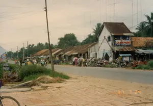 Đây là một khung cảnh hồ nước rộng lớn dưới bầu trời nhiều mây. Phía đối diện là một ngọn đồi thoai thoải được bao phủ bởi thảm cỏ xanh mướt và trồng nhiều cây cối, có khả năng là cây thông, đặc trưng của khu vực Đà Lạt. Có một vài công trình kiến trúc nằm rải rác. Sát bờ hồ phía bên trái có một ngôi nhà nhỏ một tầng, có kiến trúc giống như một thủy tạ. Xa hơn trên đồi cũng có thể thấy một vài ngôi nhà khác. Mặt hồ khá tĩnh lặng. Ở góc trên bên phải của bức ảnh, một cành cây rủ xuống. Phía bờ bên phải có một phần của một cầu tàu nhỏ hoặc đài quan sát nhô ra mặt nước. Toàn bộ khung cảnh mang lại cảm giác yên bình và tĩnh lặng, không có hoạt động rõ rệt nào của con người. Không có bất kỳ văn bản hay chữ viết nào có thể nhìn thấy.。（日本語訳）。これは曇り空の下の広大な湖の光景です。対岸は、青々とした芝生に覆われ、多くの樹木が植えられたなだらかな丘で、ダラット地方に特徴的な松の木である可能性があります。いくつかの建築物が点在しています。左側の湖岸沿いには、水上家屋のような建築様式の小さな平屋の家があります。丘のさらに奥にも、他の家がいくつか見えます。湖面はかなり静かです。写真の右上の角では、木の枝が垂れ下がっています。右側の岸には、水面に突き出た小さな桟橋または展望台の一部があります。光景全体が平和で静かな感覚をもたらし、目立った人間の活動はありません。