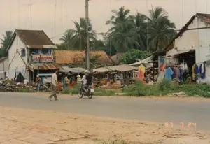 Bức ảnh ghi lại khung cảnh sinh hoạt đời thường tại một khu chợ ven đường ở một vùng quê hoặc thị trấn nhỏ của Việt Nam, vào ngày 24 tháng 7 năm 1997.。Phía bên kia đường là một khu chợ cóc nhộn nhịp, nơi người dân tụ tập mua bán dưới những mái che tạm bằng bạt. Nhiều mặt hàng được bày bán, bao gồm quần áo treo trên giá và các sản phẩm đựng trong giỏ đan. Có thể thấy một người đang đội nón lá trong khu chợ.。Phía sau khu chợ là những ngôi nhà, trong đó có một ngôi nhà hai tầng với tấm biển hiệu lớn màu đỏ trên ban công ghi rõ: "HIỆU VÀNG KIM PHỤNG" và bên dưới là dòng chữ "MUA BÁN - TRAO ĐỔI VÀNG BẠC ĐÁ QUÝ". Các ngôi nhà khác có mái ngói và một số đã xuống cấp. Phía sau cùng là những hàng cây dừa cao vút.。Trên con đường nhựa, một người đàn ông đang đi bộ và một chiếc xe máy chở hai người đang lưu thông. Dọc lề đường, nhiều chiếc xe máy khác đang đậu. Một chi tiết đặc trưng của thời kỳ này là cột điện bê tông có gắn loa truyền thanh công cộng (loa phường) và rất nhiều ăng-ten TV trên các mái nhà, phản ánh bối cảnh xã hội và công nghệ của thập niên 90.。（日本語訳）。1997年7月24日、ベトナムの田舎または小さな町の道端にある市場での日常生活の風景を記録した写真です。道の向こう側は賑やかな青空市場で、人々がシートでできた仮設の屋根の下に集まって売買をしています。ラックに掛けられた服や編みかごに入れられた製品など、多くの商品が並べられています。市場の中にはノンラーをかぶった人の姿も見られます。市場の後ろには家々があり、その中には2階建ての家が一軒あります。そのバルコニーには大きな赤い看板があり、「HIỆU VÀNG KIM PHỤNG」と明記され、その下には「MUA BÁN - TRAO ĐỔI VÀNG BẠC ĐÁ QUÝ」という文字があります。他の家は瓦屋根で、いくつかは老朽化しています。一番後ろには、高くそびえるヤシの木が立ち並んでいます。アスファルトの道路上では、一人の男性が歩いており、二人乗りのバイク一台が走っています。道端には、他のバイクが何台も駐車しています。この時代を特徴づける一つのディテールは、公共放送用のスピーカー（街区スピーカー）が取り付けられたコンクリートの電柱と、家々の屋根にある非常に多くのテレビアンテナであり、90年代の社会と技術の背景を反映しています。