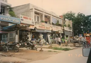 Bối cảnh là một con phố ở Việt Nam vào ngày 24 tháng 7 năm 1997, được thể hiện qua con số "97 7 24" ở góc dưới bên phải của bức ảnh.。Đây là một khu phố thương mại với một dãy nhà phố hai tầng liền kề. Con đường phía trước có một phần là đường đất và một phần đã được trải nhựa. Nhiều cửa hàng đang mở cửa kinh doanh.。Các bảng hiệu có thể đọc được từ trái sang phải bao gồm:。- Bảng hiệu màu xanh lam với chữ nổi "NGUYỄN - HOÀNG".。- Bảng hiệu màu trắng với chữ đỏ "福 PHƯỚC TÀI".。- Bảng hiệu "TƯỜNG - PHÁT".。- Các bảng hiệu ở xa hơn có thể đọc được một phần là "PHƯỚC HƯNG" và "PHƯỚC KÝ".。Hoạt động trên phố khá nhộn nhịp. Nhiều người đang di chuyển bằng xe máy (xe máy) và xe đạp (xe đạp). Có một người phụ nữ mặc áo đỏ, đội nón lá đang đạp xe. Nhiều chiếc xe máy, chủ yếu là các dòng xe cũ, được dựng trước các cửa hàng. Một số người đang đi bộ trên vỉa hè. Một cửa hàng dường như đang trưng bày các bộ phận xe máy hoặc hàng hóa tương tự trên kệ. Bức ảnh được chụp từ bên trong một chiếc ô tô, có thể thấy một phần gương chiếu hậu ở góc dưới bên phải.。（日本語訳）。背景は1997年7月24日のベトナムのある通りで、写真の右下隅にある「97 7 24」という数字で示されています。ここは、2階建てのタウンハウスが連なる商店街です。手前の道は一部が未舗装で、一部が舗装されています。多くの店が営業しています。左から右へ読み取れる看板には、以下が含まれます。- 「NGUYỄN - HOÀNG」と浮き彫りにされた青い看板。- 赤い文字で「福 PHƯỚC TÀI」と書かれた白い看板。- 「TƯỜNG - PHÁT」の看板。- 遠くにある看板は、部分的に「PHƯỚC HƯNG」と「PHƯỚC KÝ」と読み取れます。通りの様子はかなり賑やかです。多くの人がバイク（xe máy）や自転車（xe đạp）で移動しています。赤い服を着てノンラーをかぶった女性が自転車を漕いでいます。多くのバイク、主に旧型のモデルが店の前に停められています。何人かの人が歩道を歩いています。ある店は、バイクの部品かそれに類する商品を棚に陳列しているようです。写真は車内から撮影されたもので、右下隅にサイドミラーの一部が見えます。