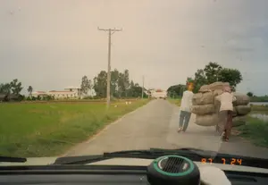 Bức ảnh được chụp từ bên trong một chiếc ô tô, ghi lại cảnh quan trên một con đường nông thôn vào ngày 24 tháng 7 năm 1997, như được hiển thị trên đồng hồ điện tử ở bảng điều khiển xe: '97 7 24'. Trên đường, hai người đang vận chuyển hàng hóa bằng một chiếc xe kéo tay. Chiếc xe chất đầy những bao tải lớn. Một người phụ nữ mặc áo hoa, quần dài và đầu quấn khăn hồng đang đẩy xe từ phía sau. Một người khác mặc áo trắng, quần tối màu và đầu quấn khăn màu cam đang đi phía trước. Hai bên đường là cánh đồng lúa xanh. Ở phía xa bên trái có một tòa nhà lớn, hai tầng, mái đỏ và một vài ngôi nhà nhỏ hơn. Dọc theo con đường có cây cối và các cột điện.。（日本語訳）。写真は車内から撮影されたもので、車のダッシュボードにあるデジタル時計に「'97 7 24」と表示されているように、1997年7月24日の田舎道の風景を捉えています。路上では、二人が手押し車で荷物を運んでいます。手押し車には大きな袋が満載です。花柄のシャツと長ズボンを着用し、頭にピンクのスカーフを巻いた女性が、後ろから手押し車を押しています。もう一人は、白いシャツと暗い色のズボンを着用し、頭にオレンジ色のスカーフを巻いて、前を歩いています。道の両側は緑の田んぼです。遠くの左側には、大きな二階建ての赤い屋根の建物と、いくつかのより小さな家があります。道に沿って木々や電柱があります。