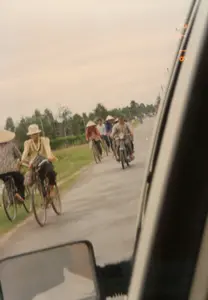 Bức ảnh chụp một cảnh giao thông trên một con đường nhựa ở vùng nông thôn Việt Nam. Khung cảnh cho thấy một nhóm người đang di chuyển cùng chiều bằng xe đạp và xe máy. Ở phía trước, có hai người phụ nữ đang đi xe đạp. Người phụ nữ bên trái mặc áo hoa và đội nón lá. Người phụ nữ bên phải mặc áo sơ mi kẻ ca rô màu vàng và đội một chiếc mũ vải. Đi sau họ là một nhóm khác, bao gồm một người đàn ông đang lái xe máy và những người khác đi xe đạp, một số cũng đội nón lá. Hai bên đường là bãi cỏ xanh và hàng cây. Bức ảnh có vẻ được chụp từ bên trong một chiếc ô tô đang chạy, vì có một phần khung cửa sổ xe ở bên phải và gương chiếu hậu ở góc dưới bên trái. Ở cạnh phải của ảnh có dấu ngày tháng kỹ thuật số màu cam, hiển thị các con số "97 7 24", cho biết ngày chụp là 24 tháng 7 năm 1997.。（日本語訳）。この写真はベトナムの田舎にあるアスファルトの道路での交通風景を撮影したものです。その風景は、自転車やバイクに乗った人々の集団が同じ方向に移動している様子を示しています。前方には、自転車に乗っている2人の女性がいます。左側の女性は花柄の服を着て、ノンラーをかぶっています。右側の女性は黄色のチェック柄のシャツを着て、布製の帽子をかぶっています。彼女たちの後ろには別の集団がおり、バイクを運転している男性1人と、自転車に乗っている他の人々が含まれ、そのうちの何人かもノンラーをかぶっています。道路の両側には緑の草地と並木があります。この写真は走行中の車内から撮影されたようです。なぜなら、右側に車の窓枠の一部が、左下隅にサイドミラーが写っているからです。写真の右端にはオレンジ色のデジタル日付表示があり、「97 7 24」という数字が表示されています。これは撮影日が1997年7月24日であることを示しています。