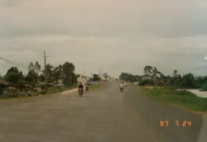 Bức ảnh chụp một con đường nhựa ở vùng nông thôn Việt Nam, dẫn đến một cây cầu lớn màu trắng ở phía xa. Bầu trời u ám, có nhiều mây. Trên đường có một vài người đang điều khiển xe máy và một người đi xe đạp. Bên trái đường là những ngôi nhà dân đơn sơ, một vài quán nhỏ lợp bạt, cùng với cây cối và cột điện. Có một tấm biển báo màu xanh lam bên đường nhưng chữ viết trên đó không thể đọc được. Bên phải đường là một dải đất cỏ xanh và một con sông hoặc kênh rạch. Khung cảnh phản ánh một ngày bình thường ở một vùng quê hoặc ngoại ô Việt Nam vào năm 1997. Dấu ngày tháng '97 7 24' được in ở góc dưới bên phải của bức ảnh.。（日本語訳）。ベトナムの田舎にあるアスファルトの道を写した写真で、遠くにある大きな白い橋へと続いています。空はどんよりとしており、雲が多いです。道路にはバイクを運転している数人と、自転車に乗っている一人がいます。道路の左側には、簡素な民家、防水シートで屋根を覆ったいくつかの小さな店、そして木々や電柱があります。道端には青い看板がありますが、そこに書かれている文字は読み取れません。道路の右側には、緑の草地と川または運河があります。この光景は、1997年におけるベトナムの田舎または郊外の日常の一日を反映しています。写真の右下隅には「'97 7 24」という日付が印字されています。