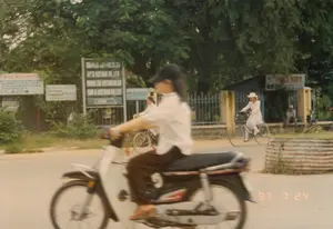 Bức ảnh chụp cảnh ven đường ở Việt Nam vào ngày 24 tháng 7 năm 1997. Ở tiền cảnh, một người đang lái một chiếc xe máy trên đường, hình ảnh bị mờ do chuyển động cho thấy xe đang chạy. Người lái xe đội mũ lưỡi trai, mặc áo sơ mi trắng và quần tối màu. Ở hậu cảnh, một phụ nữ mặc áo dài trắng và đội nón lá đang đi xe đạp. Có những người khác cũng đang đi xe đạp và một người đàn ông đang ngồi bên lề đường, gần một công trình nhỏ có hàng rào. Cảnh vật xung quanh có nhiều cây xanh. Ven đường có một số biển báo. Một biển báo lớn màu xanh lam liệt kê tên các công ty: 'SAIGON VIET-HAN CO. LTD', 'APCS VIETNAM CO. LTD', 'YOUNG JIN VIETNAM CO. LTD', 'SAM A VIETNAM CO. LTD', và một mũi tên chỉ đường ghi '11KM'. Một công trình nhỏ giống như ki-ốt có biển hiệu ghi 'THOẠI KIẾT'. Các biển báo khác bị mờ không thể đọc rõ.。（日本語訳）。1997年7月24日にベトナムで撮影された道端の風景写真。前景には、道路でバイクを運転している人がおり、その姿は動きによってブレており、バイクが走行中であることを示している。運転手は野球帽をかぶり、白いシャツと暗い色のズボンを着用している。背景には、白いアオザイを着てノンラーをかぶった女性が自転車に乗っている。他にも自転車に乗っている人々がおり、一人の男性が柵のある小さな建物の近くの道端に座っている。周囲の景色には緑の木々が多く見られる。道端にはいくつかの看板がある。大きな青い看板には、「SAIGON VIET-HAN CO. LTD」、「APCS VIETNAM CO. LTD」、「YOUNG JIN VIETNAM CO. LTD」、「SAM A VIETNAM CO. LTD」といった会社名が列挙されており、「11KM」と書かれた方向を示す矢印がある。キオスクのような小さな建物には「THOẠI KIẾT」と書かれた看板がある。他の看板はぼやけていて、はっきりと読み取ることはできない。