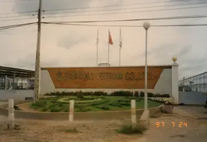 Bức ảnh chụp cổng chính của công ty Samyang Vietnam Co., Ltd. vào ngày 24 tháng 7 năm 1997, như được hiển thị bởi dấu ngày tháng màu cam ở góc dưới bên phải. Trên bức tường lớn xây bằng gạch màu cam, có dòng chữ kim loại 'SAMYANG VIETNAM CO., LTD.' cùng với logo của công ty. Phía sau bức tường là ba cột cờ, trong đó có một lá cờ màu đỏ và hai lá cờ trắng có biểu tượng. Phía trước cổng là một bồn hoa được trồng cỏ và các loại cây cảnh, tạo thành một vòng tròn trang trí. Con đường phía trước cổng là đường đất, chưa được trải nhựa, hai bên có các cọc tiêu bê tông. Bên trái có một cột điện bằng gỗ với nhiều dây điện. Xa hơn phía sau hàng rào có thể nhìn thấy một phần của các tòa nhà nhà xưởng. Bầu trời u ám, có nhiều mây.。（日本語訳）。1997年7月24日、右下隅にあるオレンジ色のタイムスタンプが示すように、Samyang Vietnam Co., Ltd.社の正門を撮影した写真。オレンジ色のレンガで建てられた大きな壁には、会社のロゴとともに「SAMYANG VIETNAM CO., LTD.」という金属製の文字がある。壁の後ろには3本の旗竿があり、そのうち1本は赤い旗、2本はシンボルマークの入った白い旗である。門の前には、草や観葉植物が植えられた花壇があり、装飾的な円を形成している。門の前の道は未舗装の土の道で、両側にはコンクリートの杭がある。左側には、多くの電線がついた木製の電柱がある。空は曇っており、雲が多い。