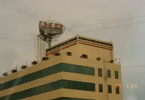 Bức ảnh chụp một tòa nhà văn phòng hiện đại, nhiều tầng vào ngày 24 tháng 7 năm 1997. Tòa nhà được sơn màu be và có các dải cửa sổ kính lớn màu xanh sẫm.。Các thông tin bằng chữ có thể đọc được bao gồm:。1. Trên nóc nhà có một biển hiệu lớn hình elip với dòng chữ "HD" màu đỏ và "BÌNH DƯƠNG" màu đen, được đặt trên một kết cấu giàn thép.。2. Trên mặt tiền của tầng trên cùng, có dòng chữ tiếng Việt màu sẫm: "HIỆP HỘI SẢN XUẤT KINH DOANH HÀNG XUẤT NHẬP KHẨU VÀ PHÁT TRIỂN CÁC NGÀNH NGHỀ TỈNH BÌNH DƯƠNG".。3. Ngay bên dưới dòng chữ tiếng Việt là một dòng chữ tiếng Anh màu đỏ, có vẻ là bản dịch, một số từ khó đọc nhưng có thể đoán là "BINH DUONG ASSOCIATION OF ENTERPRISES FOR INVESTMENT & DEVELOPMENT".。Về khung cảnh và các vật thể:。- Địa điểm: Đây là một tòa nhà văn phòng, có thể là trụ sở của một hiệp hội doanh nghiệp ở khu vực đô thị tại tỉnh Bình Dương.。- Vật thể: Tòa nhà, biển hiệu, dây điện, thang sắt bên ngoài. Sân thượng được trang trí bằng nhiều lá cờ nhỏ nhiều màu và các chậu cây cảnh.。- Hoạt động: Không có hoạt động cụ thể nào của con người được ghi lại. Khung cảnh tĩnh lặng.。（日本語訳）。1997年7月24日に撮影された、現代的な多層階のオフィスビルの写真です。建物はベージュ色に塗られており、濃い青色の大きなガラス窓の帯があります。読み取れる文字情報は以下の通りです：。1. 屋上には、鉄骨のトラス構造の上に設置された、赤い文字で「HD」、黒い文字で「BÌNH DƯƠNG」と書かれた大きな楕円形の看板があります。2. 最上階のファサードには、濃い色のベトナム語で「HIỆP HỘI SẢN XUẤT KINH DOANH HÀNG XUẤT NHẬP KHẨU VÀ PHÁT TRIỂN CÁC NGÀNH NGHỀ TỈNH BÌNH DƯƠNG」という文字があります。3. ベトナム語の文字のすぐ下には、赤い英語の文字があり、それは翻訳文のようです。いくつかの単語は読みにくいですが、「BINH DUONG ASSOCIATION OF ENTERPRISES FOR INVESTMENT & DEVELOPMENT」と推測できます。風景と物体について：。- 場所：これはオフィスビルで、ビンズオン省の都市部にある企業協会の本部である可能性があります。- 物体：建物、看板、電線、外部の鉄製はしご。屋上は、多くの色とりどりの小さな旗や観葉植物の鉢で飾られています。- 活動：人間の具体的な活動は記録されていません。静かな光景です。