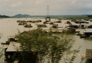 Trên ảnh có dấu ngày tháng '97 7 24'. Bức ảnh ghi lại khung cảnh một làng nổi trên một vùng nước rộng lớn, có thể là một hồ chứa hoặc một khúc sông lớn. Hàng chục ngôi nhà nổi, hay còn gọi là nhà bè, được dựng san sát nhau. Các ngôi nhà này có kết cấu đơn giản, chủ yếu làm bằng gỗ, một số lợp mái tôn, một số khác lợp mái lá. Đây có thể là một làng bè nuôi cá, nơi người dân sinh sống và nuôi trồng thủy sản trực tiếp dưới nhà của họ. Nhiều chiếc thuyền, ghe nhỏ đậu cạnh các nhà bè, là phương tiện di chuyển chính. Một điểm đặc biệt nổi bật trong khung cảnh là một trụ điện cao thế lớn được xây dựng ngay giữa mặt nước. Ở phía xa là đường chân trời với những ngọn đồi và cây cối ven bờ. Phần tiền cảnh của bức ảnh bị nhòe bởi một lùm cây, cho thấy ảnh được chụp khi đang di chuyển.。（日本語訳）。写真には「'97 7 24」という日付スタンプがあります。この写真は、貯水池か大きな川の一部分かもしれない広大な水域に浮かぶ水上村の風景を捉えています。何十もの水上家屋、別名いかだ家屋が、互いに密集して建てられています。これらの家は単純な構造で、主に木でできており、一部はトタン屋根、その他は茅葺き屋根です。これは魚の養殖いかだ村である可能性があり、人々は自分たちの家の真下で生活し、水産養殖を行っています。多くの小舟やボートがいかだ家屋の隣に停泊しており、主要な移動手段となっています。風景の中で特に目立つ点は、水面の真ん中に建てられた大きな高圧電線の鉄塔です。遠くには、丘と岸辺の木々がある地平線が見えます。写真の前景部分は木の茂みによってぼやけており、移動中に撮影されたことを示しています。