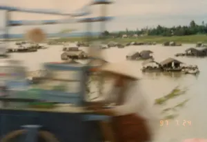 Góc dưới bên phải của ảnh có dấu ngày tháng '97 7 24'. Bức ảnh bị mờ, có thể được chụp từ một phương tiện đang di chuyển, ghi lại khung cảnh một làng nổi trên một vùng nước rộng lớn như hồ hoặc sông. Trên mặt nước có rất nhiều nhà nổi, hay còn gọi là nhà bè, được làm bằng vật liệu đơn giản. Một vài nhà bè có thuyền nhỏ neo đậu bên cạnh. Những ngôi nhà này là nơi sinh sống của người dân, có khả năng liên quan đến hoạt động nuôi trồng thủy sản hoặc đánh bắt cá. Ở phía xa là đường bờ với cây cối xanh tươi. Do ảnh bị mờ, không thể xác định rõ các hoạt động cụ thể đang diễn ra.。（日本語訳）。写真の右下隅には「'97 7 24」という日付スタンプがあります。写真はぼやけており、移動中の乗り物から撮影された可能性があり、湖や川のような広大な水域に浮かぶ水上集落の光景を捉えています。水面には、いかだハウスとも呼ばれる、簡素な材料で作られた多くの水上家屋があります。いくつかのいかだハウスの隣には、小さなボートが停泊しています。これらの家は人々の住居であり、水産養殖や漁業活動に関連している可能性があります。遠くには、緑豊かな木々が生い茂る岸辺が見えます。写真がぼやけているため、行われている具体的な活動を明確に特定することはできません。