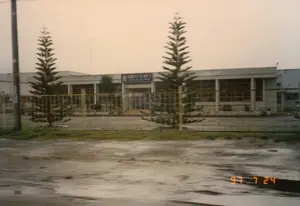 Bức ảnh chụp mặt tiền của một tòa nhà một tầng, có khả năng là một nhà máy hoặc văn phòng công ty. Trên lối vào chính có một tấm biển lớn màu xanh với dòng chữ: 'CÔNG TY TƠ VIỆT-Ý' và bên dưới là 'VIETNAM - ITALY SILK COMPANY'. Tòa nhà được rào lại bằng hàng rào lưới kim loại. Phía trước sân có trồng hai cây tùng. Trên mái nhà có thể nhìn thấy các cột ăng-ten. Cảnh vật phía trước, bao gồm mặt đường có vẻ ẩm ướt và lầy lội, bị nhòe, cho thấy bức ảnh được chụp từ một phương tiện đang di chuyển. Bầu trời u ám, tạo cảm giác đây là một ngày sau cơn mưa. Ở góc dưới bên phải của ảnh có dấu ngày kỹ thuật số màu cam '97 7 24'.。（日本語訳）。写真は、工場または会社の事務所と思われる1階建ての建物の正面を撮影したものです。正面の入り口の上には、「CÔNG TY TƠ VIỆT-Ý」と書かれ、その下に「VIETNAM - ITALY SILK COMPANY」と書かれた大きな青い看板があります。建物は金属製のメッシュフェンスで囲まれています。前庭には2本の松の木が植えられています。屋根の上にはアンテナの柱が見えます。空は曇っており、雨上がりの日であるという印象を与えます。写真の右下隅には、「97 7 24」というオレンジ色のデジタル日付スタンプがあります。