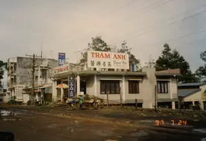 Hình ảnh ghi lại một khung cảnh đường phố ở Bảo Lộc vào ngày 24 tháng 7 năm 1997. Bầu trời u ám, mặt đường là đường đất lầy lội và có vũng nước, cho thấy trời có thể vừa mưa.。Nổi bật trong ảnh là một cửa hàng tên là "TRÂM ANH", chuyên kinh doanh trà và cà phê. Có nhiều bảng hiệu được nhìn thấy:。- Bảng hiệu lớn trên nóc nhà ghi "TRÂM ANH", chữ Hán "簪纓名茶" (Danh Trà - Trà nổi tiếng), địa chỉ "807 TRẦN PHÚ, BẢO LỘC" và số điện thoại "TEL: 063.864403".。- Bảng hiệu ở góc tòa nhà ghi "TRÀ CÀ PHÊ TRÂM ANH".。- Bảng hiệu dọc màu xanh ghi "trà TRÂM ANH".。- Một bảng hiệu khác ở phía sau ghi "TRÀ Café BLAO", trong đó B'Lao là tên cũ của Bảo Lộc.。Về hoạt động, có một người đang đội nón lá đứng cạnh hai chiếc xe đạp. Các xe đạp này dường như được chất đầy hàng hóa. Phía trước cửa hàng có một chiếc ô nhiều màu. Vài chiếc xe máy cũng đang đậu gần đó. Khung cảnh đặc trưng của Việt Nam thời kỳ này với những ngôi nhà bê tông, hệ thống dây điện chằng chịt trên các cột điện.。（日本語訳）。1997年7月24日のバオロクの街の風景を記録した写真です。空はどんよりとしており、路面はぬかるんだ土の道で水たまりができており、雨が降ったばかりかもしれないことを示しています。写真の中で目立っているのは「TRÂM ANH」という名前の店で、お茶とコーヒーを専門に扱っています。多くの看板が見られます：。- 屋根の上の大きな看板には、「TRÂM ANH」、漢字の「簪纓名茶」（Danh Trà - 有名な茶）、住所「807 TRẦN PHÚ, BẢO LỘC」と電話番号「TEL: 063.864403」が記されています。- 建物の角にある看板には「TRÀ CÀ PHÊ TRÂM ANH」と書かれています。- 青い縦長の看板には「trà TRÂM ANH」と書かれています。- 後方にある別の看板には「TRÀ Café BLAO」と書かれており、B'Laoはバオロクの旧名です。人々の様子については、ノンラーをかぶった一人の人物が2台の自転車のそばに立っています。これらの自転車は荷物でいっぱいになっているようです。店の前にはカラフルな傘が1本あります。数台のバイクも近くに停まっています。コンクリートの家々、電柱に複雑に張り巡らされた電線といった、この時代のベトナムに特徴的な光景です。