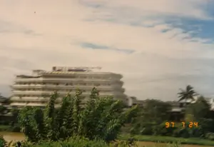 Bức ảnh chụp một khung cảnh ven sông hoặc khu vực có nhiều cây cối, nhìn về phía một tòa nhà lớn, nhiều tầng. Tòa nhà này có màu trắng, kiến trúc có phần uốn lượn với nhiều cửa sổ. Trên nóc tòa nhà có một biển hiệu lớn với các chữ màu đỏ, nhưng do ảnh mờ nên không thể đọc rõ nội dung. Phía trước, gần với người chụp là những lùm cây xanh tươi rậm rạp, hình ảnh hơi nhòe cho thấy có thể ảnh được chụp khi đang di chuyển. Bên trái có thể thấy một phần của một con sông hoặc kênh với nước màu nâu. Ở phía xa hơn, bên phải, có nhiều cây xanh, trong đó có một cây dừa. Bầu trời có mây trắng và xanh. Dấu ngày tháng "97 7 24" được in màu vàng ở góc dưới bên phải của bức ảnh.。（日本語訳）。写真は、川沿いまたは木々が多い地域の風景を写しており、大きな多層階の建物の方を向いている。この建物は白色で、多くの窓があり、やや曲線的な建築様式である。建物の屋上には赤い文字の大きな看板があるが、写真がぼやけているため、その内容ははっきりと読み取れない。手前、撮影者に近い位置には、青々とした生い茂る木々があり、画像が少しぶれていることから、移動中に撮影された可能性があることがわかる。さらに遠くの右側には、多くの緑の木々があり、その中にはヤシの木が1本ある。空には白い雲と青空が広がっている。写真の右下隅に「97 7 24」という日付が黄色で印字されている。