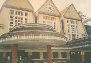 Bức ảnh chụp mặt tiền của Ga Đà Lạt, một công trình kiến trúc cổ. Trên phần mái nhà trung tâm có dòng chữ "DALAT" màu đỏ. Phía trên dòng chữ là một chiếc đồng hồ lớn. Tòa nhà có màu vàng, với ba mái nhà hình tam giác lợp ngói. Kiến trúc nổi bật với nhiều cửa sổ kính màu. Một chiếc loa phát thanh được gắn ở phần mái nhà bên trái. Lối vào chính có một mái che hình tròn lớn được chống đỡ bởi các cột trụ. Dưới mái che có thể thấy bóng dáng của một vài người, có thể là hành khách hoặc nhân viên nhà ga. Dấu ngày tháng kỹ thuật số màu cam ở góc dưới bên phải ghi rõ "97 7 25", cho biết bức ảnh được chụp vào ngày 25 tháng 7 năm 1997.。（日本語訳）。ダラット駅の正面を写した写真で、古い建築物である。中央の屋根部分には、赤い「DALAT」の文字がある。文字の上には大きな時計がある。建物は黄色で、瓦で葺かれた3つの三角屋根がある。建築は多くのステンドグラスの窓で際立っている。左側の屋根部分には放送用のスピーカーが取り付けられている。正面玄関には、柱で支えられた大きな円形の庇がある。庇の下には数人の人影が見え、乗客か駅員であろう。右下のオレンジ色のデジタル日付表示には「97 7 25」とはっきりと記されており、写真が1997年7月25日に撮影されたことを示している。