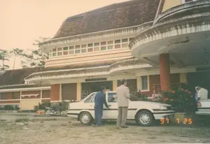 Bức ảnh chụp bên ngoài một tòa nhà lớn có kiến trúc cổ, được xác định là Ga xe lửa Đà Lạt qua biển hiệu và phong cách kiến trúc đặc trưng. Trên lối vào chính của tòa nhà có một biển hiệu ghi rõ dòng chữ "CỬA VÀO GA".。Địa điểm là một nhà ga xe lửa với kiến trúc kiểu Pháp, có mái ngói màu nâu, các bức tường màu vàng và đỏ, cùng dãy cửa sổ kính nhiều màu. Phía trước nhà ga là một khoảng sân đất rộng, chưa được lát nền, có cỏ mọc thưa thớt. Hoạt động chính diễn ra là có hai người đàn ông mặc trang phục lịch sự (vest) đang đứng cạnh một chiếc ô tô sedan màu trắng. Một người đàn ông mặc vest xanh đang mở cửa phía người lái. Người còn lại đứng bên cạnh. Ở phía xa bên phải, có một người đàn ông khác đang đứng gần một chiếc xe hơi khác. Bên trong cửa ra vào có thể thấy lờ mờ vài người. Các vật thể trong ảnh bao gồm tòa nhà ga, chiếc ô tô sedan màu trắng, một chiếc xe máy kiểu cũ đậu ở phía sau bên trái, các bụi hoa màu đỏ được trồng ven tòa nhà và những cây thông ở phía hậu cảnh.。（日本語訳）。写真は、看板と特徴的な建築様式からダラット駅と特定される、古い建築様式の大きな建物の外で撮影されたものです。建物の正面玄関の上には、「CỬA VÀO GA」（駅入口）という文字がはっきりと書かれた看板があります。場所はフランス風建築の駅で、茶色の瓦屋根、黄色と赤色の壁、そして色とりどりのガラス窓が並んでいます。駅の前には舗装されていない広い土の庭があり、草がまばらに生えています。主な活動として、きちんとした服装（ベスト）の男性二人が白いセダン車の隣に立っています。青いベストを着た男性が運転席側のドアを開けています。もう一人はその隣に立っています。右奥には、別の男性が別の車の近くに立っています。出入口の内側には、数人がぼんやりと見えます。写真の中の物体には、駅舎、白いセダン車、左後方に停まっている旧式のバイク、建物の縁に植えられた赤い花の茂み、そして背景の松の木が含まれます。