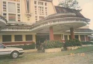 Bức ảnh chụp mặt tiền của một tòa nhà lớn, có khả năng là một nhà hàng hoặc một công trình công cộng ở Đà Lạt, được chụp vào ngày 25 tháng 7 năm 1997. Tòa nhà có kiến trúc kiểu cũ, có thể là từ thời Pháp thuộc, với tường sơn màu vàng và nâu đỏ, đã có dấu hiệu của thời gian. Điểm nhấn là mái hiên lớn, uốn lượn che khu vực lối vào và được chống đỡ bởi các cột tròn. Mái nhà được lợp ngói. Phía trên lối vào chính có một tấm biển hiệu nhưng chữ viết đã mờ và không thể đọc được. Tương tự, phía trên một cửa sổ ở tầng hai cũng có chữ nhưng không rõ. Khung cảnh tĩnh lặng, không có hoạt động nào của con người đang diễn ra. Phía trước tòa nhà là một khoảng sân đất và cỏ, nơi có một chiếc ô tô sedan màu bạc đời cũ và một chiếc xe máy đang đậu. Dọc theo mặt tiền là các bồn cây lớn trồng hoa dâm bụt màu đỏ. Bên trong một cửa sổ ở tầng trên có thể thấy một bức chân dung lớn đã bị phai màu.。（日本語訳）。1997年7月25日に撮影された、ダラットにある大きな建物、おそらくレストランか公共施設の正面を写した写真です。建物は古い様式の建築で、おそらくフランス植民地時代のものであり、壁は黄色と赤褐色に塗られていますが、経年劣化の兆候が見られます。特徴的なのは、入口エリアを覆う大きな波形の庇で、円柱によって支えられています。屋根は瓦で葺かれています。正面玄関の上には看板がありますが、文字は薄れていて読むことができません。同様に、2階の窓の上にも文字がありますが、はっきりしません。人の活動は見られず、静かな光景です。建物の前には土と草の庭があり、そこには古い型のシルバーのセダン車1台とバイク1台が停まっています。建物の正面に沿って、赤いハイビスカスの花が植えられた大きなプランターが並んでいます。上階の窓の内側には、色褪せた大きな肖像画が見えます。