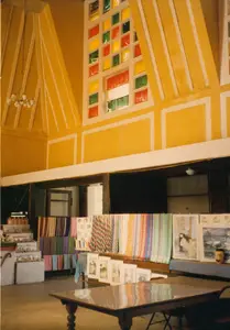 Bức ảnh chụp bên trong một tòa nhà có kiến trúc đặc biệt, có thể là một cửa hàng lưu niệm hoặc một điểm dừng chân cho khách du lịch ở khu vực Đà Lạt. Nổi bật là phần trần nhà cao, được sơn màu vàng với các dầm trang trí màu trắng tạo thành các đường nét góc cạnh. Phía trên cao có một ô cửa sổ lớn được ghép từ nhiều ô kính màu sặc sỡ (đỏ, vàng, xanh lá).。Không có hoạt động nào của con người diễn ra, chỉ có một không gian trưng bày hàng hóa. Các mặt hàng được bày bán bao gồm nhiều loại vải và khăn choàng dệt với màu sắc và hoa văn đa dạng (sọc, ziczac) được treo trên giá. Bên cạnh đó là các tác phẩm nghệ thuật như tranh vẽ một cô gái tóc dài, tranh phong cảnh và một số bản in nhỏ hơn được đặt trên kệ. Ở phía trước, có một chiếc bàn gỗ lớn với mặt bàn được đánh bóng. Phía bên trái có các kệ bậc thang trưng bày các món đồ nhỏ hơn. Một chiếc đèn chùm kiểu cũ treo trên trần nhà.。（日本語訳）。特徴的な建築様式の建物内部を撮影した写真で、ダラット地方の土産物店か、観光客向けの休憩所かもしれません。目立つのは高い天井で、黄色に塗られ、白い装飾的な梁が角張った線を描いています。高い位置には、色とりどりのガラス（赤、黄、緑）を多数組み合わせて作られた大きな窓があります。人の活動は見られず、商品を展示する空間だけがあります。販売されている商品には、多様な色や模様（ストライプ、ジグザグ）の様々な種類の織物やショールが含まれ、ラックに掛けられています。その隣には、長い髪の少女を描いた絵、風景画、そしていくつかの小さな版画などの芸術作品が棚に置かれています。手前には、天板が磨かれた大きな木製のテーブルがあります。左側には、より小さな品々を展示する階段状の棚があります。古風なシャンデリアが天井から吊り下げられています。