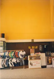 Bức ảnh chụp lại khung cảnh bên trong Ga Đà Lạt vào năm 1997. Trên bức tường lớn màu nâu ở trung tâm có dòng chữ "GA ĐÀ LẠT" màu vàng. Đây là một không gian có vẻ như là phòng chờ hoặc một cửa hàng tiện lợi bên trong nhà ga.。Bên trái là một giá treo bằng gỗ bày bán nhiều loại quần áo, chủ yếu là áo khoác và áo gi-lê nhiều màu sắc, có thể là đồ lưu niệm.。Bên phải là một quầy hàng nhỏ phục vụ giải khát. Mặt trước của quầy có các dòng chữ "COFFEE BOSS", "COLD DRINK", "KARAOKE". Trên quầy và trong tủ kính có trưng bày nhiều loại đồ uống như nước ngọt đóng lon, nước suối và rượu. Cạnh đó là một chiếc tủ lạnh màu trắng, một chiếc tivi kiểu cũ và một tấm áp phích quảng cáo.。Phía trên biển hiệu "GA ĐÀ LẠT" có một chiếc đồng hồ treo tường. Toàn bộ không gian có tường sơn màu vàng và sàn nhà có rãnh. Khung cảnh cho thấy một hoạt động kinh doanh phục vụ hành khách và khách du lịch tại nhà ga.。（日本語訳）。1997年のダラット駅内部の光景を写した写真です。中央にある大きな茶色の壁には、「GA ĐÀ LẠT」という黄色い文字があります。ここは駅の待合室か、あるいは駅構内の売店のような空間です。左側には木製の陳列棚があり、主にお土産品と思われる色とりどりのジャケットやベストなど、様々な衣類が販売されています。右側には、飲み物を提供する小さなカウンターがあります。カウンターの正面には「COFFEE BOSS」、「COLD DRINK」、「KARAOKE」という文字が見えます。カウンターの上とガラスケースの中には、缶ジュース、ミネラルウォーター、お酒といった様々な飲み物が陳列されています。その隣には、白い冷蔵庫、旧式のテレビ、そして広告ポスターが置かれています。「GA ĐÀ LẠT」の看板の上には、壁掛け時計があります。空間全体は、黄色く塗られた壁と溝のある床が特徴です。この光景は、駅で乗客や観光客にサービスを提供する商業活動の様子を示しています。