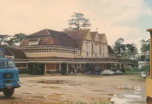 Bức ảnh chụp quang cảnh phía trước Ga Đà Lạt vào ngày 25 tháng 7 năm 1997. Tòa nhà ga là một công trình kiến trúc cổ độc đáo, sơn màu vàng với mái ngói màu nâu. Trên phần mái trung tâm có ba chóp nhọn, một chiếc đồng hồ và lá cờ Việt Nam. Dòng chữ 'ĐÀ LẠT' được ghi trên tường, ngay phía trên cửa sổ chính. Sân ga là một bãi đất rộng, lầy lội và có nhiều vũng nước, cho thấy trời vừa mưa. Ở bên trái, một chiếc xe tải cũ màu xanh lam hiệu IFA đang đỗ. Phía trước hiên nhà ga có hai chiếc ô tô con và một vài bộ bàn ghế nhựa màu xanh, nơi có vài người đang ngồi và đứng, có thể đang chờ đợi hoặc nghỉ ngơi. Xung quanh là cây cối, chủ yếu là thông, đặc trưng của Đà Lạt. Ở góc phải của ảnh, có một phần của một vật thể màu vàng, có thể là một toa tàu hoặc thiết bị đường sắt.。（日本語訳）。1997年7月25日のダラット駅前の光景を撮影した写真です。駅舎はユニークな古い建築物で、黄色に塗られ、茶色の瓦屋根が特徴です。中央の屋根部分には、3つの尖塔、1つの時計、そしてベトナムの国旗があります。「ĐÀ LẠT」という文字が、中央の窓のすぐ上の壁に書かれています。駅前広場は広々とした地面で、ぬかるんでおり水たまりが多く、雨が降ったばかりであることがわかります。左側には、IFAブランドの古い青いトラックが1台駐車しています。駅舎の軒先には、2台の乗用車といくつかの青いプラスチック製のテーブルと椅子のセットがあり、数人が座ったり立ったりしています。おそらく待っているか、休憩しているのでしょう。周囲には木々があり、そのほとんどがダラット特有の松です。写真の右隅には、黄色い物体の一部が見えますが、それは客車か鉄道設備かもしれません。