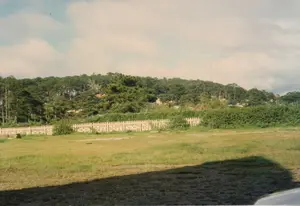 Đây là một khung cảnh thiên nhiên ở vùng cao nguyên, có khả năng là ở Đà Lạt. Tiền cảnh là một bãi đất rộng có cỏ mọc, với một bóng râm lớn đổ xuống, cho thấy bức ảnh có thể được chụp từ một nơi có mái che. Ở góc dưới cùng bên phải, có thể thấy một phần nhỏ của một chiếc ô tô. Ở giữa khung cảnh là một hàng rào cọc gỗ màu trắng. Trên hàng rào có vẻ như có chữ viết hoặc hình vẽ nhưng không thể đọc rõ nội dung. Phía sau hàng rào là các bụi cây xanh tươi. Hậu cảnh là một ngọn đồi được bao phủ bởi rừng thông rậm rạp, một cảnh quan đặc trưng của Đà Lạt. Xa xa phía trên ngọn đồi có thể thấy một cột tháp truyền thông hoặc ăng-ten. Bầu trời trong xanh và có mây trắng.。（日本語訳）。これは高原地帯の自然風景で、おそらくダラットでしょう。前景には草が生えた広い地面があり、大きな影が落ちています。これは写真が屋根のある場所から撮影された可能性を示唆しています。右下隅には、自動車の一部が小さく見えます。風景の中央には、白い木製の杭の柵があります。柵の上には文字か絵が描かれているようですが、内容ははっきりと読み取れません。柵の後ろには青々とした茂みがあります。背景は、ダラット特有の景観である、鬱蒼とした松林に覆われた丘です。丘の上の遠くには、通信塔かアンテナが見えます。空は青く澄んでおり、白い雲があります。