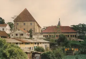 Bức ảnh ghi lại một quang cảnh khu dân cư trên sườn đồi tại Đà Lạt, được chụp vào ngày 25 tháng 7 năm 1997, thể hiện qua dấu ngày tháng "97 7 25" ở góc dưới bên phải. Khung cảnh cho thấy nhiều công trình kiến trúc đa dạng. Ở phía sau, nổi bật là một tòa nhà lớn bằng đá với mái ngói hình chóp cao, bên cạnh là một công trình khác có mái ngói đỏ và một tháp chuông nhỏ, gợi nhớ đến kiến trúc của một nhà thờ hoặc tu viện. Ở tiền cảnh và trung cảnh là những ngôi nhà dân được xây dựng san sát nhau với các loại mái khác nhau, bao gồm mái ngói và mái tôn đã gỉ sét. Cây cối xanh tươi, đặc biệt là cây thông, bao quanh khu vực. Phía trước có một mảnh vườn rau nhỏ. Không có hoạt động nào rõ rệt ngoài cảnh sinh hoạt thường ngày như quần áo phơi trên ban công.。（日本語訳）。この写真は、1997年7月25日に撮影されたダラットの丘の中腹にある住宅地の光景を記録したもので、右下の「97 7 25」という日付表示で示されています。この風景は、多くの多様な建築物を示しています。後方には、高い円錐形の瓦屋根を持つ大きな石造りの建物が際立っており、その隣には赤い瓦屋根と小さな鐘楼を持つ別の建物があり、教会や修道院の建築を彷彿とさせます。前景と中景には、瓦屋根や錆びたトタン屋根など、さまざまな種類の屋根を持つ民家が密集して建てられています。緑豊かな木々、特に松の木が、この地域を囲んでいます。手前には小さな菜園があります。バルコニーに干された洗濯物のような日常の生活風景以外に、目立った活動は見られません。