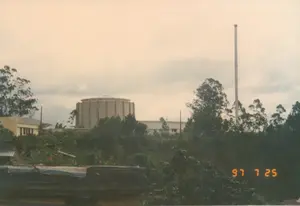 Trên ảnh có dấu ngày tháng màu cam '97 7 25'.。Bức ảnh chụp một quang cảnh nhìn từ xa về phía một khu phức hợp công trình, được xác định là Viện Nghiên cứu Hạt nhân Đà Lạt.。Địa điểm: Một khu vực có nhiều cây cối ở Đà Lạt, nhìn ra một cơ sở nghiên cứu khoa học.。Các vật thể và công trình:。- Tòa nhà chính: Một công trình kiến trúc lớn, hình trụ tròn bằng bê tông, là tòa nhà chứa lò phản ứng hạt nhân.。- Ống khói: Một ống khói công nghiệp cao, thẳng đứng nằm ở bên phải tòa nhà chính.。- Các tòa nhà phụ: Các tòa nhà thấp hơn với mái bằng nằm xung quanh tòa nhà chính.。- Cây cối: Rất nhiều cây xanh rậm rạp, bao gồm cả cây cao và bụi cây, bao quanh các công trình. Ở phía trước có một khu vườn nhỏ với vài bông hoa màu cam.。- Tiền cảnh: Một phần của một cấu trúc không xác định, có thể là mái nhà hoặc bờ tường, được che phủ bởi một tấm bạt.。Hoạt động: Bức ảnh là một cảnh quan tĩnh, không có hoạt động nào của con người được ghi lại một cách rõ ràng. Bầu trời nhiều mây.。（日本語訳）。写真にはオレンジ色の日付スタンプ「'97 7 25」があります。この写真は、ダラット原子力研究所と特定される複合施設を遠くから見た風景を撮影したものです。場所：ダラットの木々が多い地域で、科学研究施設が見えます。物体と建造物：。- 主要な建物：コンクリート製の大きな円筒形の建築物で、原子炉を収容する建物です。- 煙突：主要な建物の右側にある、高くて垂直な工業用の煙突。- 付属の建物：主要な建物の周りにある、平らな屋根を持つ低い建物群。- 樹木：高木や低木を含む、たくさんの青々とした木々が建物を囲んでいます。手前には、オレンジ色の花がいくつか咲いている小さな庭があります。- 前景：屋根か壁の一部と思われる、防水シートで覆われた不特定の構造物の一部。活動：写真は静的な風景であり、人間の活動は明確には記録されていません。空は曇っています。