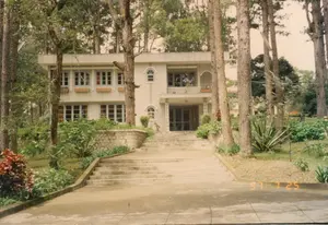 Bức ảnh chụp một tòa nhà hai tầng, có thể là một biệt thự hoặc nhà khách, nằm giữa một khu rừng thông rậm rạp, một khung cảnh đặc trưng của Đà Lạt. Một lối đi và cầu thang rộng bằng bê tông dẫn lên sảnh vào chính của tòa nhà. Tòa nhà có kiến trúc khá hiện đại với mặt tiền màu sáng, nhiều cửa sổ kính và một ban công ở tầng hai. Xung quanh tòa nhà là một khu vườn với nhiều loại cây cối và cây cảnh, được bao quanh bởi những bức tường đá thấp. Rất nhiều cây thông cao lớn bao bọc lấy công trình, tạo nên một không gian yên tĩnh và biệt lập. Không có hoạt động nào của con người được ghi lại. Ở góc dưới bên phải bức ảnh có dấu ngày tháng '97 7 25', cho biết ảnh được chụp vào ngày 25 tháng 7 năm 1997.。（日本語訳）。写真は、ダラット特有の風景である、鬱蒼とした松林の中に建つ、別荘かゲストハウスと思われる2階建ての建物を写している。コンクリート製の広い通路と階段が、建物のメインエントランスホールへと続いている。建物はかなり現代的な建築様式で、明るい色のファサード、多くのガラス窓、そして2階にバルコニーがある。建物の周りには、様々な樹木や観葉植物が植えられた庭があり、低い石垣で囲まれている。多くの背の高い松の木が建物を包み込み、静かで隔離された空間を作り出している。人の活動は記録されていない。写真の右下隅には「'97 7 25」という日付印があり、写真が1997年7月25日に撮影されたことを示している。