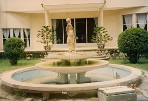 Bức ảnh chụp quang cảnh sân trước của một tòa nhà, có thể là một biệt thự hoặc nhà nghỉ. Ở trung tâm là một đài phun nước nhiều tầng bằng đá nhưng không hoạt động, nước trong bể tù đọng và có rêu. Trên đỉnh đài phun nước có một bức tượng tạc hình một người phụ nữ theo phong cách cổ điển. Phía sau là tòa nhà với mặt tiền màu sáng, có cửa kính lớn và rèm cửa màu trắng. Hai bên hiên nhà đặt hai chậu cây tùng cảnh trong chậu gốm sứ trang trí, trên một chậu có thể thấy một ký tự Hán tự (có thể là chữ 'Thọ'). Xung quanh sân còn có các bụi cây được cắt tỉa hình tròn và bãi cỏ. Không có hoạt động nào của con người được ghi lại. Trên ảnh có dấu ngày tháng "97 7 25".。（日本語訳）。ある建物の前庭の光景を写した写真で、別荘かペンションの可能性があります。中央には石造りの多層式噴水がありますが稼働しておらず、水盤の水はよどんで苔が生えています。噴水の頂上には、古典的なスタイルの女性をかたどった彫像があります。後方には、明るい色のファサードを持つ建物があり、大きなガラスのドアと白いカーテンがあります。玄関の両脇には、装飾的な陶磁器の鉢に植えられた松の盆栽が2つ置かれており、片方の鉢には漢字（おそらく「寿」の字）が見えます。庭の周りには、丸く刈り込まれた植え込みや芝生もあります。人の活動は記録されていません。写真には「97 7 25」という日付スタンプがあります。