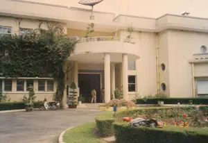 Bức ảnh chụp bên ngoài một tòa nhà lớn, có kiến trúc kiểu biệt thự ở Đà Lạt, có khả năng là một dinh thự hoặc nhà khách. Tòa nhà hai tầng sơn màu kem, nổi bật với mái hiên hình tròn có các cột trụ chống đỡ và một ban công cong ở tầng trên. Trên mái nhà có lắp một chảo vệ tinh, một vật dụng tương đối hiện đại vào thời điểm năm 1997. Một phần mặt tiền bên trái được bao phủ bởi giàn hoa giấy lớn có hoa màu đỏ.。Khuôn viên phía trước có một khu vườn được chăm sóc kỹ lưỡng với các hàng rào cây được cắt tỉa, luống hoa hồng và cây cảnh trong chậu. Một chiếc xe máy, có vẻ là loại Honda Cub, đang đậu ở sân trước. Tại lối vào chính, có hai người đàn ông đang đứng. Ở tiền cảnh, trên một hàng rào cây, có một vài chiếc áo khoác và túi xách. Không có văn bản nào có thể đọc được, nhưng dấu ngày tháng '97 7 25' hiển thị ở góc dưới bên phải.。（日本語訳）。写真は、ダラットにあるヴィラ風の建築様式の大きな建物の外観を写したもので、邸宅またはゲストハウスである可能性があります。建物はクリーム色に塗られた2階建てで、支柱で支えられた円形の庇と、上階の湾曲したバルコニーが特徴的です。屋根の上にはパラボラアンテナが設置されており、1997年当時にしては比較的新しいものです。ファサードの左側の一部は、赤い花を咲かせた大きなブーゲンビリアの棚で覆われています。前庭には、刈り込まれた生垣、バラの花壇、鉢植えの観葉植物がある、手入れの行き届いた庭園があります。ホンダ・カブと思われるバイクが1台、前庭に停まっています。正面玄関には、2人の男性が立っています。前景の生垣の上には、いくつかのジャケットとバッグが置かれています。読み取れる文字はありませんが、右下隅に「'97 7 25」という日付スタンプが表示されています。