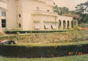 Bức ảnh ghi lại khung cảnh một khu vườn hoa rộng lớn phía trước một dinh thự mang phong cách kiến trúc Art Deco, có thể là một biệt thự nghỉ dưỡng ở Đà Lạt. Tòa nhà chính có màu vàng kem, nổi bật với các cửa sổ hình tròn và hình chữ nhật có mái hiên che nắng. Một dãy hành lang có mái vòm chạy dọc một phần của công trình.。Khu vườn được thiết kế theo kiểu bậc thang, bao gồm các luống hoa hồng đang nở rộ với nhiều màu sắc như đỏ, hồng và trắng. Xung quanh các luống hoa là những hàng rào cây được cắt tỉa vuông vắn, tạo nên một bố cục ngăn nắp. Phía trước là một bãi cỏ xanh mướt. Ở góc trái, trên một hàng rào cây, có vẻ như là một chiếc áo khoác hoặc túi xách bị bỏ lại. Phía sau dinh thự là những cây thông cao vút, một hình ảnh quen thuộc của vùng cao nguyên Đà Lạt.。Không có hoạt động nào của con người được ghi lại, khung cảnh rất tĩnh lặng và yên bình. Ở góc dưới cùng bên phải của hình ảnh có một dấu ngày kỹ thuật số màu cam hiển thị "97 7 25", cho biết thời điểm chụp là ngày 25 tháng 7 năm 1997.。（日本語訳）。この写真は、アールデコ様式の邸宅の前にある広大な花畑の風景を捉えており、おそらくダラットの別荘であろう。主要な建物はクリームイエローで、日よけのある円形および長方形の窓が特徴的である。アーチ型の屋根を持つ廊下が、建物の一部に沿って続いている。庭園は段々畑式に設計されており、赤、ピンク、白など様々な色の満開のバラの花壇がある。花壇の周りには、四角く刈り込まれた生け垣があり、整然とした配置を作り出している。手前には青々とした芝生が広がっている。邸宅の後ろには、ダラット高原の馴染み深い光景である、高くそびえる松の木々がある。人の活動は記録されておらず、風景は非常に静かで穏やかである。画像の右下隅には、「97 7 25」と表示されたオレンジ色のデジタル日付印があり、撮影日時が1997年7月25日であることを示している。