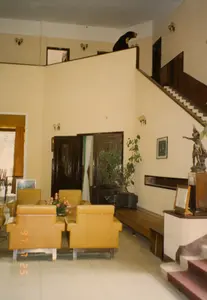 Đây là hình ảnh chụp bên trong sảnh chờ của một tòa nhà, có thể là khách sạn hoặc nhà nghỉ vào năm 1997. Không gian được bài trí với một bộ ghế sofa bọc da màu vàng mù tạt. Phía sau là cầu thang gỗ dẫn lên tầng trên, nơi có một con gấu nhồi bông được đặt trên lan can như một vật trang trí. Các vật dụng khác trong phòng bao gồm một chậu cây cảnh, một chiếc tivi kiểu cũ (CRT), một bức tượng đồng đặt trên bệ, và các đèn tường. Trên ảnh có dấu ngày tháng "97 7 25". Có một văn bản trong khung đặt trên bệ gần cầu thang nhưng không thể đọc rõ nội dung. Không có người hay hoạt động nào đang diễn ra trong ảnh.。（日本語訳）。これは1997年のある建物、おそらくホテルか旅館の待合ロビーの内部を撮影した写真です。空間にはマスタードイエローの革張りソファセットが置かれています。後ろには上の階へ続く木製の階段があり、その手すりには装飾としてテディベアが置かれています。部屋にあるその他の物には、観葉植物の鉢、旧式のテレビ（ブラウン管）、台座に置かれた銅像、そして壁の照明が含まれます。写真には「97 7 25」という日付のスタンプがあります。階段の近くの台座に置かれた額縁の中に文章がありますが、内容ははっきりと読み取れません。写真には人や進行中の活動はありません。