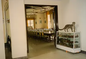 Bức ảnh chụp bên trong một tòa nhà, có vẻ là một nhà hàng hoặc nhà khách. Nhìn qua khung cửa gỗ tối màu là một phòng ăn hoặc phòng họp rộng rãi. Trong phòng, một chiếc bàn dài bằng gỗ được sắp xếp cùng nhiều chiếc ghế có đệm ngồi màu vàng và bọc vải trắng ở lưng ghế. Căn phòng được chiếu sáng bởi các cụm đèn trần và có cửa sổ với rèm màu vàng. Trên tường có treo một vài bức tranh trong khung. Ở phía tiền cảnh, bên phải, có một tủ kính trưng bày có bánh xe. Bên trong tủ chứa nhiều đồ vật nhỏ, có thể là đồ lưu niệm hoặc đồ trang trí. Đáng chú ý nhất là trên nóc tủ có một con vật nhồi bông, trông giống một con báo gấm. Sàn nhà được lát gạch men. Không có người trong ảnh, cho thấy không gian đang không được sử dụng. Ở góc dưới bên phải có dấu ngày tháng màu cam '97 7 25'.。（日本語訳）。写真は建物の中で撮影されたもので、レストランかゲストハウスのようです。部屋の中には、長い木製のテーブルが、座面が黄色いクッションで背もたれが白い布で覆われた多くの椅子と共に配置されています。部屋は複数の天井照明で照らされており、黄色いカーテンのかかった窓があります。壁には額に入った絵が数枚掛けられています。前景の右側には、キャスター付きのガラス製陳列棚があります。棚の中には多くの小物が収められており、お土産か装飾品だと思われます。最も注目すべきは、棚の上にウンピョウに似た動物のぬいぐるみがあることです。床はセラミックタイルで敷き詰められています。写真には人が写っておらず、その空間が現在使用されていないことを示しています。右下隅にはオレンジ色の日付スタンプ「'97 7 25」があります。