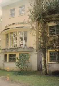 Trong ảnh có dấu ngày tháng màu cam ở góc dưới bên trái: "97 7 25".。Bối cảnh là không gian ngoài trời, chụp mặt tiền của một tòa nhà nhiều tầng. Dựa vào bối cảnh địa lý và kiến trúc, đây có thể là một biệt thự hoặc nhà nghỉ mang phong cách kiến trúc Pháp cổ ở Đà Lạt. Tòa nhà được sơn màu vàng, với phần chân tường tầng trệt được làm bằng đá hoặc bê tông thô. Một đặc điểm kiến trúc đáng chú ý là dãy cửa sổ kính hình vòng cung ở tầng trên. Các cửa sổ ở tầng trệt được lắp song sắt bảo vệ. Một bên tường của tòa nhà có một cây leo lớn bám vào, cành lá xum xuê. Phía trước tòa nhà là một bãi cỏ nhỏ và một cây bụi. Không có người hay hoạt động nào được ghi lại, khung cảnh tĩnh lặng.。（日本語訳）。写真の左下隅にオレンジ色の「97 7 25」という日付スタンプがあります。地理的背景と建築様式から、これはダラットにある古いフランス建築様式の別荘またはペンションである可能性があります。建物は黄色に塗られており、1階の壁の基礎部分は石または打ちっぱなしのコンクリートでできています。注目すべき建築上の特徴は、上階にあるアーチ型のガラス窓の列です。1階の窓には防犯用の鉄格子が取り付けられています。建物の片側の壁には、大きなつる植物が絡みつき、枝葉が青々と茂っています。建物の前には、小さな芝生と一本の低木があります。人や活動は記録されておらず、静かな光景です。