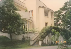 Trong ảnh có dấu ngày tháng '97 7 25'. Bức ảnh chụp một ngôi biệt thự nhiều tầng có kiến trúc hiện đại với các đường cong, có thể mang ảnh hưởng của kiến trúc Pháp. Ngôi nhà được sơn màu kem, có các ban công hình vòng cung và cửa sổ lớn. Biệt thự này nằm trên một sườn dốc, được bao quanh bởi cây cối xanh tươi, bao gồm các cây lớn giống như cây thông và nhiều bụi cây, cây leo mọc trên tường chắn bằng đá. Một cầu thang bê tông nhỏ dẫn từ con đường trải nhựa ở phía dưới lên khu vực sân vườn của ngôi nhà. Không có người xuất hiện trong ảnh, khung cảnh rất yên tĩnh, gợi liên tưởng đến một khu nghỉ dưỡng hoặc nhà ở tại Đà Lạt.。（日本語訳）。写真には「'97 7 25」という日付スタンプがあります。写真には、曲線を用いた現代的な建築様式の多層階建ての別荘が写っており、フランス建築の影響を受けている可能性があります。その家はクリーム色に塗られており、アーチ型のバルコニーと大きな窓があります。この別荘は坂の中腹にあり、松のような大きな木々や、石の擁壁に生える多くの低木やつる植物など、青々とした木々に囲まれています。下の舗装された道路から家の庭エリアへと続く小さなコンクリートの階段があります。写真には人物は写っておらず、景色は非常に静かで、ダラットのリゾート地や住宅を連想させます。