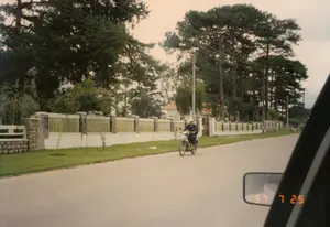 Trên ảnh có dấu ngày tháng kỹ thuật số '97 7 25'.。Đây là quang cảnh một con đường trải nhựa ở một khu vực dân cư, có khả năng là tại Đà Lạt, được chụp từ bên trong một chiếc ô tô. Điều này được thể hiện qua sự xuất hiện của gương chiếu hậu và khung cửa sổ xe ở phía bên phải bức ảnh.。Một người đàn ông đang điều khiển một chiếc xe máy trên đường. Dọc bên trái đường là một hàng rào dài, kiên cố bao quanh một khu nhà. Hàng rào này có các trụ đá và phần tường trắng. Phía sau hàng rào là một bãi cỏ xanh và một ngôi biệt thự với mái ngói màu cam ẩn mình sau những tán cây.。Khung cảnh nổi bật với rất nhiều cây thông cao lớn, đặc trưng của Đà Lạt. Các cột điện và đường dây điện cũng chạy dọc theo con đường. Ở phía xa, có thể thấy một vài người đang đi bộ.。（日本語訳）。写真には「'97 7 25」というデジタル日付スタンプがあります。これは、おそらくダラットの住宅街にあるアスファルト舗装された道路の光景で、車内から撮影されたものです。これは、写真の右側にあるバックミラーと車の窓枠の存在によって示されています。一人の男性が道路でバイクを運転しています。道路の左側には、ある敷地を囲む長くて頑丈な塀があります。この塀には石の柱と白い壁の部分があります。塀の後ろには緑の芝生と、木々の後ろに隠れたオレンジ色の瓦屋根の別荘があります。この光景は、ダラット特有のたくさんの背の高い松の木で際立っています。電柱と電線も道路に沿って走っています。遠くには、歩いている数人の人々が見えます。