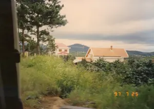 Góc dưới bên phải của hình ảnh có dấu ngày tháng màu cam: '97 7 25'. Bức ảnh chụp một khung cảnh vùng đồi núi, có thể là ở khu vực ngoại ô Đà Lạt, được nhìn từ bên trong một phương tiện đang di chuyển. Tiền cảnh là bãi cỏ dại và cây bụi um tùm. Ở trung tâm, có hai ngôi nhà kiểu biệt thự. Một ngôi nhà hai tầng với mái ngói đỏ và một ngôi nhà lớn hơn có tường trắng và mái ngói màu cam, mang kiến trúc đặc trưng của vùng. Bên trái là những cây thông cao. Phía xa là những ngọn đồi và bầu trời u ám, nhiều mây. Không có hoạt động nào của con người được thể hiện rõ, đây là một cảnh quan tĩnh lặng.。（日本語訳）。画像の右下隅には、オレンジ色の「'97 7 25」という日付スタンプがあります。この写真は、移動中の乗り物の中から見た、おそらくダラット郊外の丘陵地帯の風景を撮影したものです。前景には雑草と生い茂る低木があります。中央には、別荘風の家が2軒あります。1軒は赤い瓦屋根の2階建ての家で、もう1軒は白い壁とオレンジ色の瓦屋根を持つより大きな家で、この地域特有の建築様式をしています。左側には高い松の木があります。遠くには丘と、どんよりとした曇り空があります。人間の活動ははっきりと見られず、静かな風景です。