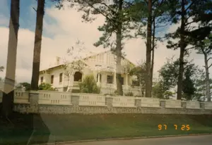 Bức ảnh chụp một biệt thự lớn màu trắng mang phong cách kiến trúc thuộc địa Pháp, tọa lạc trên một sườn đồi ở Đà Lạt. Biệt thự được bao quanh bởi một bức tường rào kiên cố, có phần móng bằng đá và phần trên bằng bê tông màu kem với các ô trống trang trí. Xung quanh biệt thự có rất nhiều cây thông cao lớn, đặc trưng của khu vực này. Phía trước là một dốc cỏ xanh mướt dẫn xuống một con đường nhựa. Bầu trời trong xanh, có vài gợn mây. Khung cảnh tĩnh lặng, không có hoạt động nào của con người được ghi lại. Ở góc dưới bên phải bức ảnh có dấu ngày tháng màu vàng ghi '97 7 25'.。（日本語訳）。写真は、ダラットの丘の中腹に位置する、フランス植民地時代の建築様式を持つ白い大きな別荘を写したものです。別荘は頑丈な塀で囲まれており、その基礎部分は石で、上部は装飾的な開口部のあるクリーム色のコンクリートでできています。別荘の周りには、この地域の特徴である背の高い松の木がたくさんあります。前面には、アスファルトの道へと続く青々とした草の斜面があります。空は青く澄んでおり、いくつかの薄雲があります。景色は静かで、人間の活動は記録されていません。写真の右下隅には、「'97 7 25」と記された黄色い日付スタンプがあります。