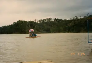 Bức ảnh được chụp vào ngày 25 tháng 7 năm 1997, ghi lại một cảnh du ngoạn trên hồ ở khu vực Đà Lạt.。Địa điểm là một hồ nước rộng, mặt nước có màu hơi đục và gợn sóng. Bối cảnh phía sau là một ngọn đồi được bao phủ bởi rừng cây rậm rạp, chủ yếu là cây thông, một cảnh quan đặc trưng của vùng cao nguyên. Bầu trời trong ảnh u ám và nhiều mây.。Hoạt động chính đang diễn ra là du ngoạn trên hồ. Ở giữa hồ, có một chiếc thuyền du lịch nhỏ, có thể là thuyền đạp nước, với hai phao màu cam và thân thuyền màu xanh. Trên thuyền có hai người đang ngồi dưới một chiếc ô sọc màu hồng và xanh để che nắng mưa. Một chiếc phao cứu sinh màu cam được treo ở bên hông thuyền. Ở góc dưới bên phải và góc dưới bên trái của bức ảnh, có thể thấy một phần của một chiếc thuyền khác hoặc một cầu tàu, cho thấy bức ảnh được chụp từ một chiếc thuyền khác hoặc từ bến.。Trên ảnh có dấu ngày tháng '97 7 25'. Không có văn bản nào khác như biển hiệu hay chữ viết trên bia.。（日本語訳）。この写真は1997年7月25日に撮影されたもので、ダラット地方の湖での遊覧風景を記録しています。場所は広い湖で、水面は少し濁った色をしており、波立っています。背景は、主に松の木からなる鬱蒼とした森に覆われた丘で、高原地帯特有の景観です。写真の空はどんよりとしており、雲が多いです。行われている主な活動は湖での遊覧です。湖の中央には、小さな遊覧船があります。おそらく足漕ぎボートで、オレンジ色のフロートが2つと青い船体が付いています。ボートの上には2人が、日雨を避けるためにピンクと青のストライプの傘の下に座っています。オレンジ色の救命浮き輪がボートの側面に掛けられています。写真の右下と左下の角には、別のボートの一部または桟橋が見え、この写真が別のボートまたは船着き場から撮影されたことを示しています。写真には「'97 7 25」という日付のスタンプがあります。看板や碑文のような他の文字はありません。