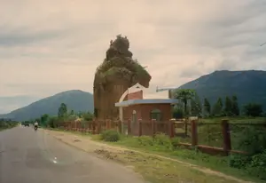 Bức ảnh chụp một di tích kiến trúc cổ, có khả năng là một Tháp Chàm, nằm ven một con đường nhựa ở vùng nông thôn. Ngọn tháp bằng gạch đã bị phong hóa nặng, phần đỉnh bị cây cỏ mọc um tùm. Bên cạnh ngọn tháp là một ngôi nhà nhỏ một tầng, có kiến trúc hiện đại hơn, được sơn màu nâu đỏ và có hàng rào bao quanh. Công trình này có thể là một trạm gác hoặc văn phòng quản lý di tích. Trên con đường, một người đang đi xe đạp và ở phía xa có một chiếc xe tải đang di chuyển. Phía sau là khung cảnh núi non trập trùng dưới bầu trời nhiều mây. Không có chữ viết nào có thể đọc được trong ảnh.。（日本語訳）。レンガ造りの塔はひどく風化しており、頂上部分は草木が生い茂っています。塔の隣には、より近代的な建築様式の小さな平屋の家があり、赤褐色に塗られ、柵で囲まれています。この建物は、警備所または遺跡の管理事務所である可能性があります。道路では、一人が自転車に乗っており、遠くにはトラックが走っています。背景には、曇り空の下に連なる山々の風景が広がっています。写真には読み取れる文字はありません。